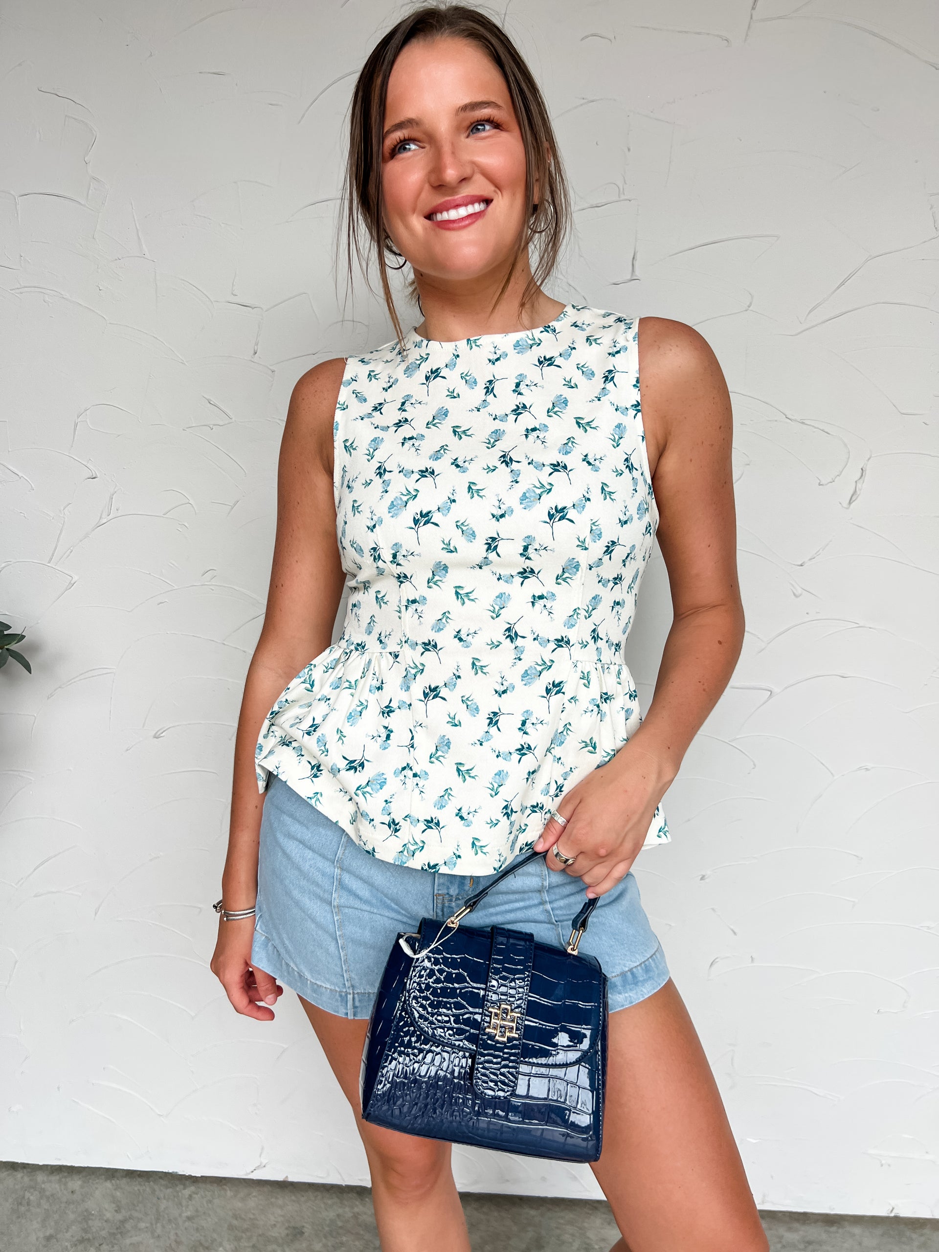 Woman wearing a floral sleeveless top and denim shorts, holding a blue handbag against a white wall.