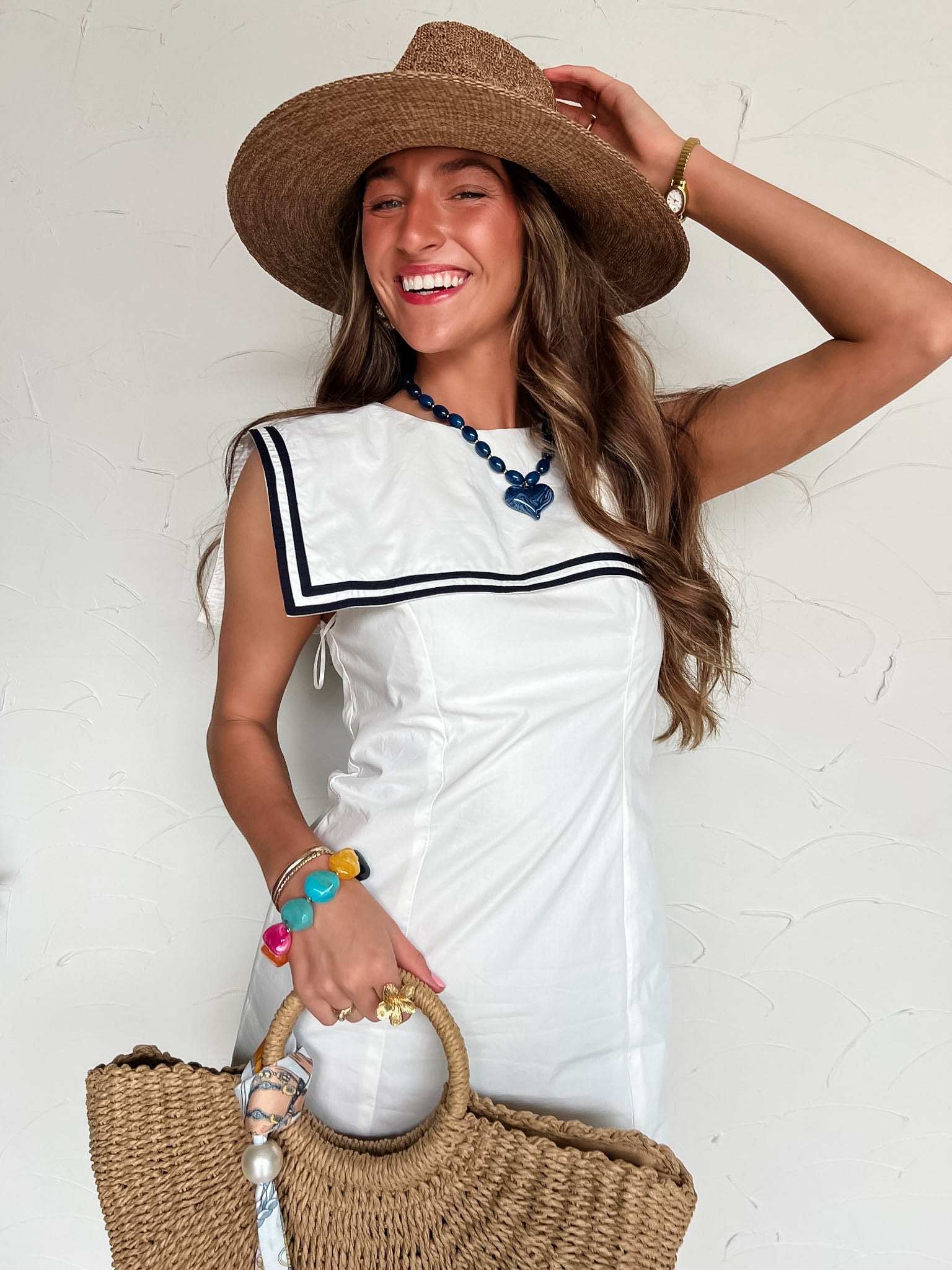 Woman in a white dress with navy detail on square collar with a straw hat and bag against a white background