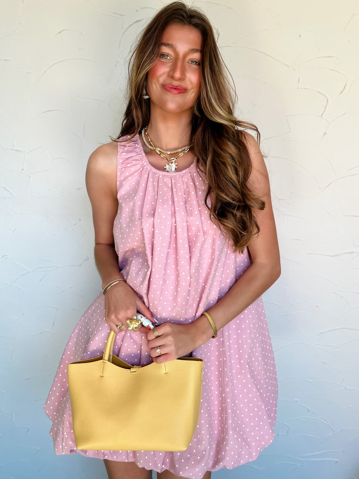 Woman wearing a pink polka dot dress holding a yellow bag against a light blue wall.