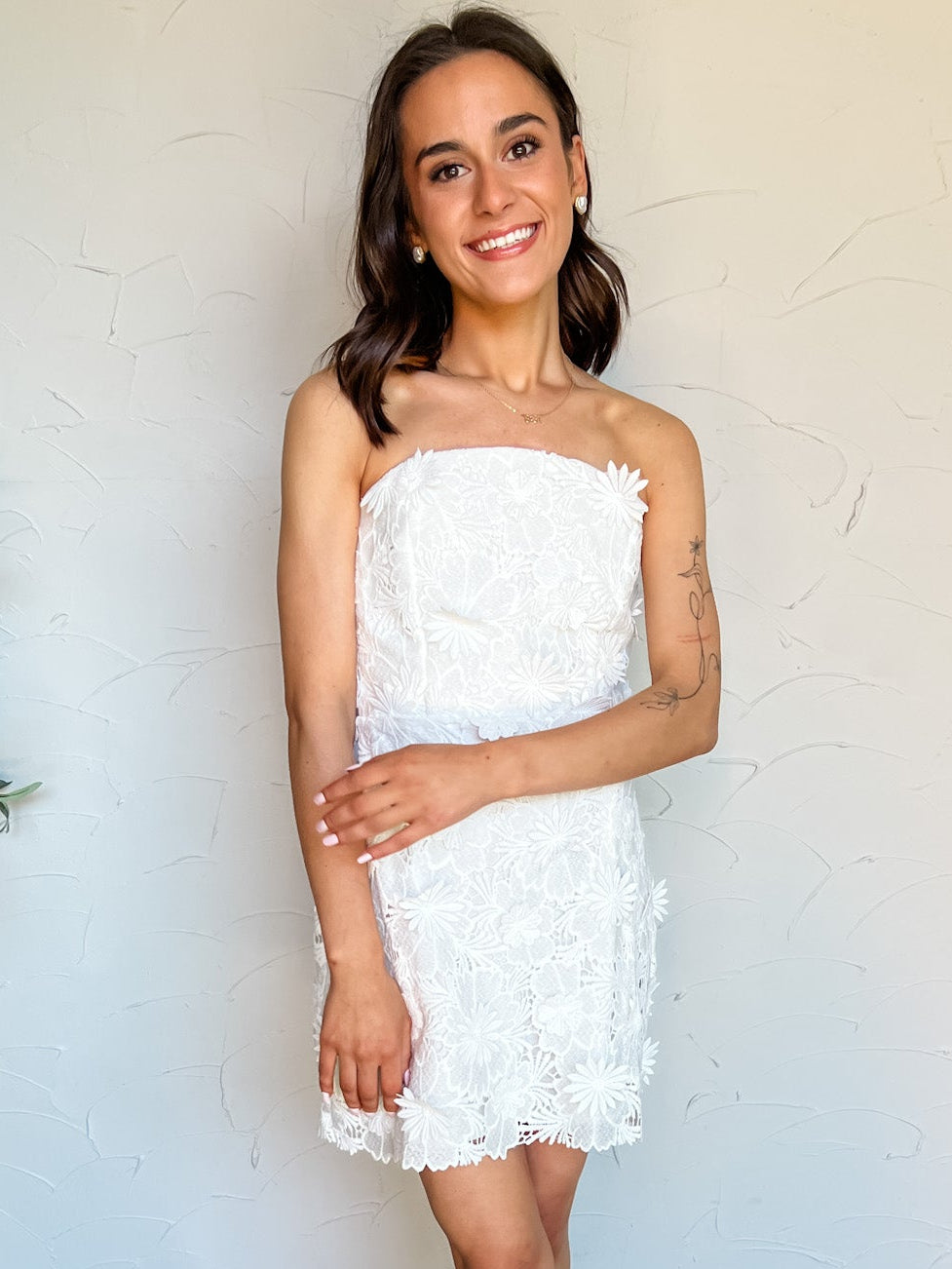 Woman wearing a white strapless lace dress against a plain wall.