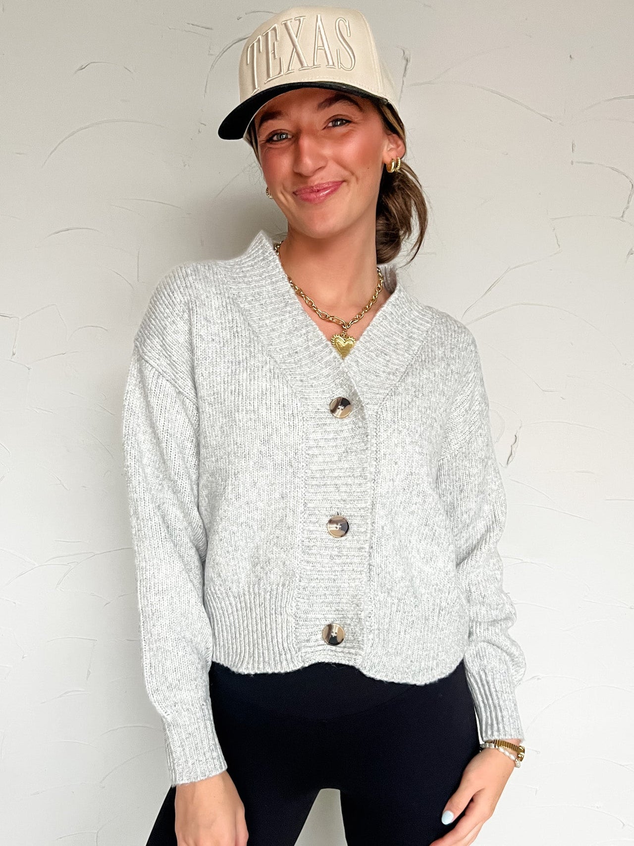 Woman wearing a light gray cardigan, black pants, and a beige cap against a white background