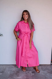 A full-length photograph of a smiling woman with straight blonde hair and bangs posing playfully with one leg lifted and one hand in her pocket against a textured white wall. She is wearing a short-sleeved tiered mini dress featuring a black and cream paisley print with bright red trim along the neckline and hem, made from an 85% rayon and 15% nylon blend fabric, styled with bright red Mary Jane flats, a black chain-strap purse, and gold bangles.