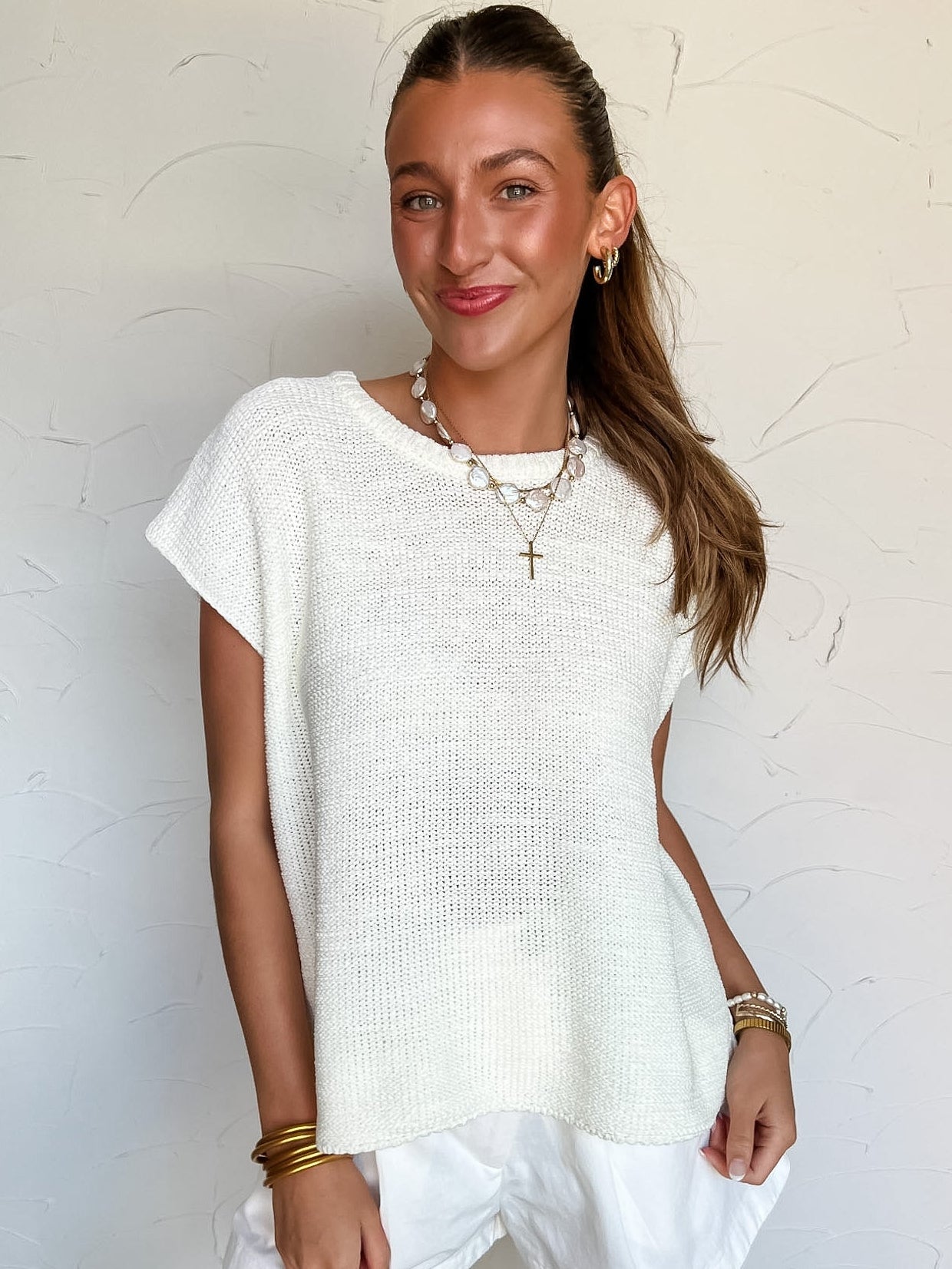 Woman wearing a white knit top and white shorts against a textured white wall.
