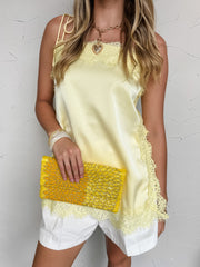 A woman standing against a textured off-white wall, wearing a pastel yellow camisole top crafted from a 100% polyester satin fabrication. The top features a straight neckline with delicate, adjustable tie-shoulder straps, a relaxed silhouette, and an asymmetrical hemline. It is beautifully detailed with eyelash lace trim along the neckline, side panels, and hem. She is styled with high-waisted white cotton shorts, a gold chain necklace with a heart pendant, a chunky acrylic bangle, and is holding a vibrant 