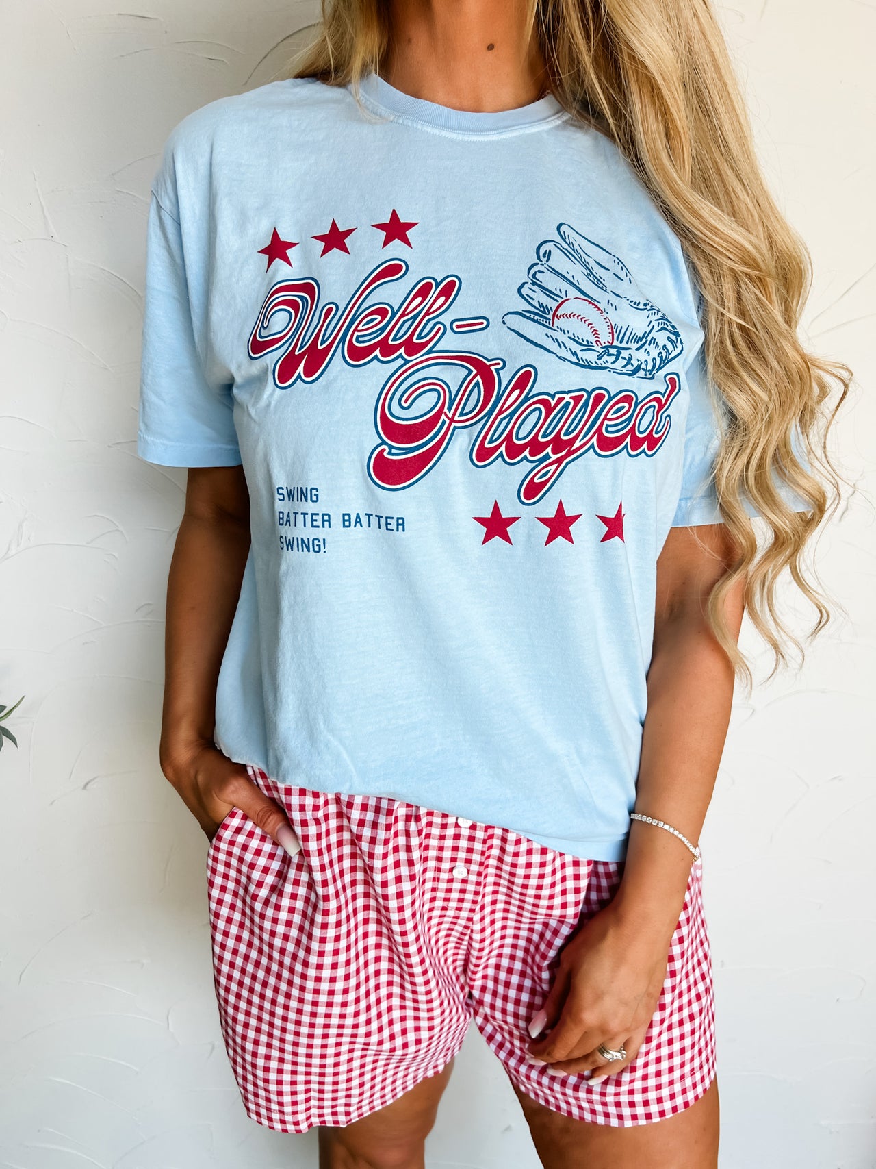 Person wearing a light blue t-shirt with red text and red and white checkered shorts on a plain background