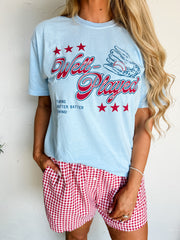Person wearing a light blue t-shirt with red text and red and white checkered shorts on a plain background