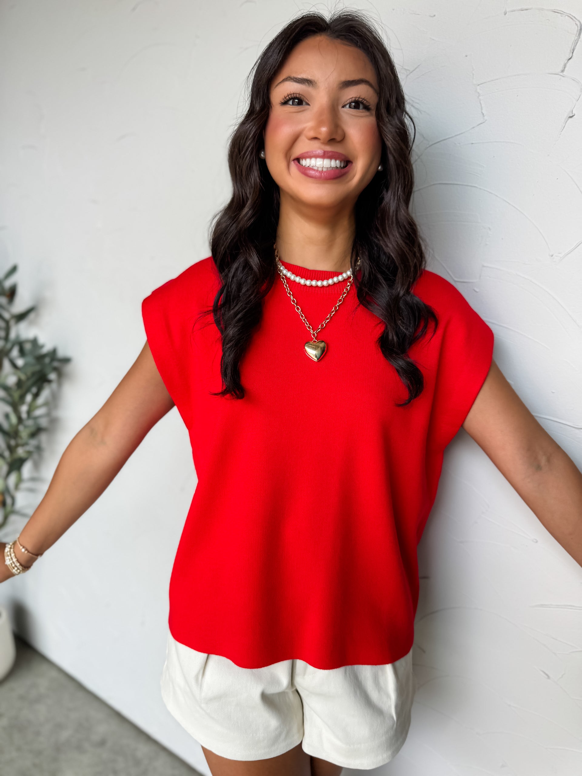 A mid-shot photograph of a smiling brunette woman posing against a textured white wall. She is wearing an oversized crewneck knit top made from a soft 80% viscose and 20% nylon blend fabric in a vibrant red colorway, featuring a fine sweater knit texture and sleeveless cap shoulders.