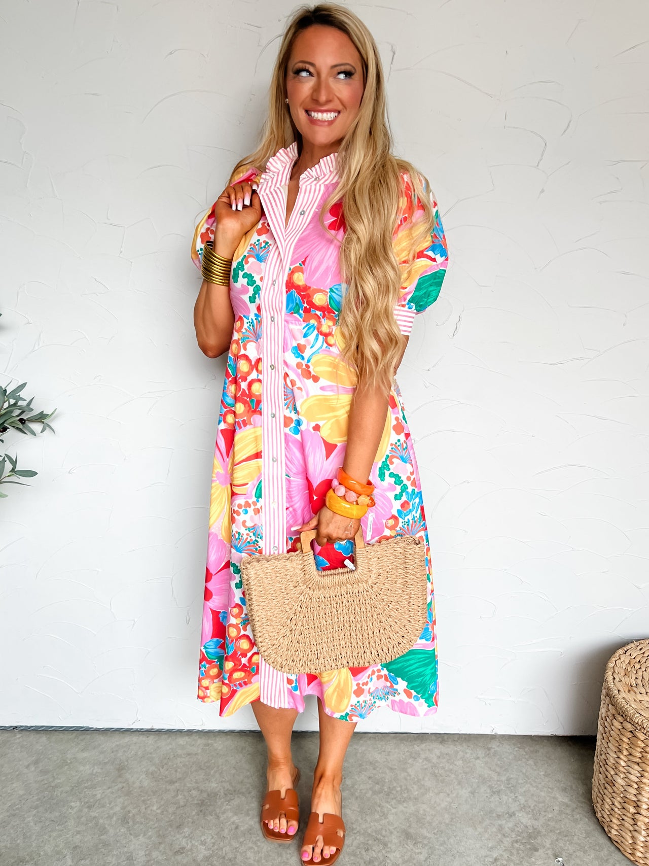 Woman wearing a pink colorful floral dress  holding a straw bag against a white background.