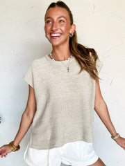 Woman wearing a beige sleeveless knit top and white shorts against a white brick wall.