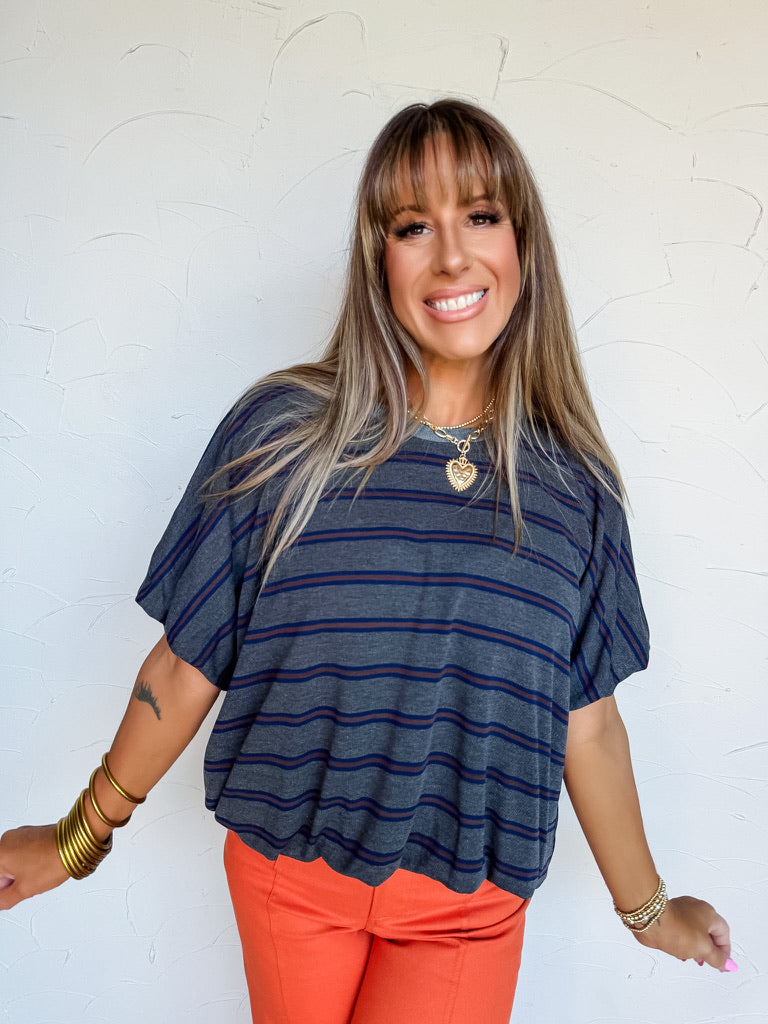 A waist-up photograph of a smiling woman with straight light-brown hair and front bangs posing with arms slightly outstretched against a textured white wall. She is wearing a short-sleeved charcoal grey knit tee made from a 40% polyester and 30% lyocell blend, featuring horizontal stripes in navy blue and rust brown, a crew neck, and wide sleeves, paired with bright orange pants and styled with stacked gold bangles, a gold heart pendant chain necklace, and a gold beaded bracelet.