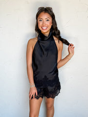 A full-length photograph of a smiling brunette woman with long dark wavy hair touching her hair while posing against a textured off-white wall next to a potted olive plant. She is wearing a black two-piece satin set made from 100% polyester, featuring a sleeveless high-neck draped halter top with black eyelash lace trim along the hem and matching black satin shorts with eyelash lace hemline detailing, styled with black-and-white zebra print loafer mules with gold horse-bit buckles, sunglasses resting on her