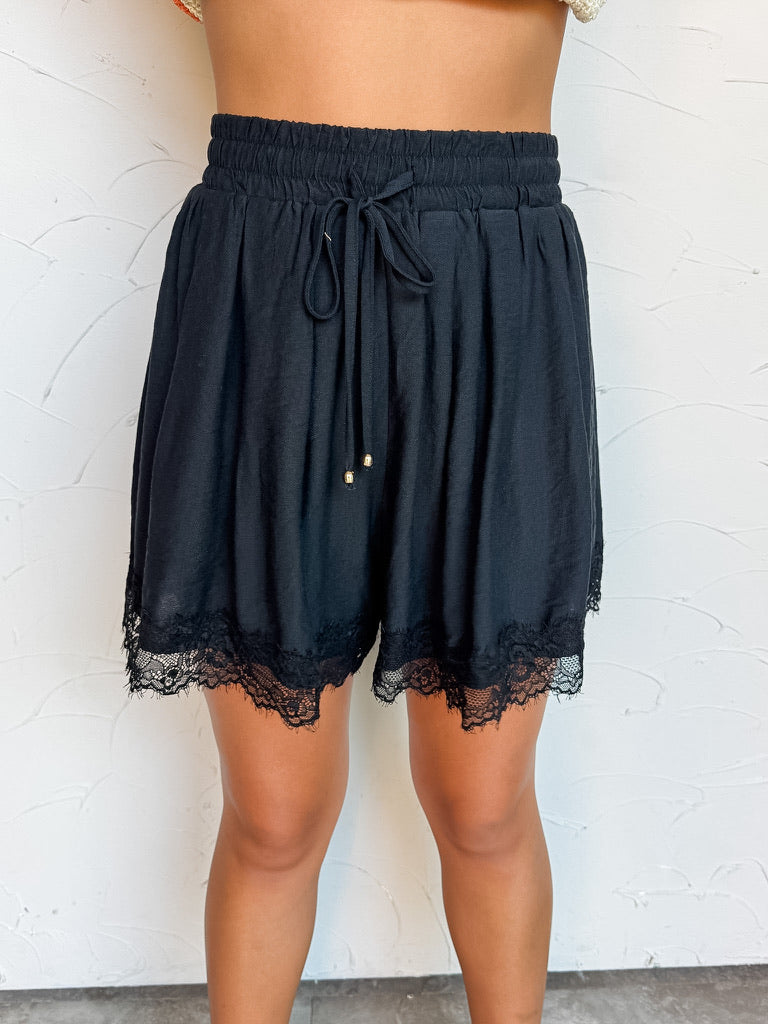 A close-up photograph from the waist down of a model wearing high-waisted solid black shorts against a textured white wall. The shorts are made from an 83% rayon and 17% nylon blend, featuring a smocked elastic waistband with a front tie drawstring detailed with gold end beads, side pockets, and delicate black eyelash lace trimming the bottom hem.