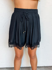 A close-up photograph from the waist down of a model wearing high-waisted solid black shorts against a textured white wall. The shorts are made from an 83% rayon and 17% nylon blend, featuring a smocked elastic waistband with a front tie drawstring detailed with gold end beads, side pockets, and delicate black eyelash lace trimming the bottom hem.