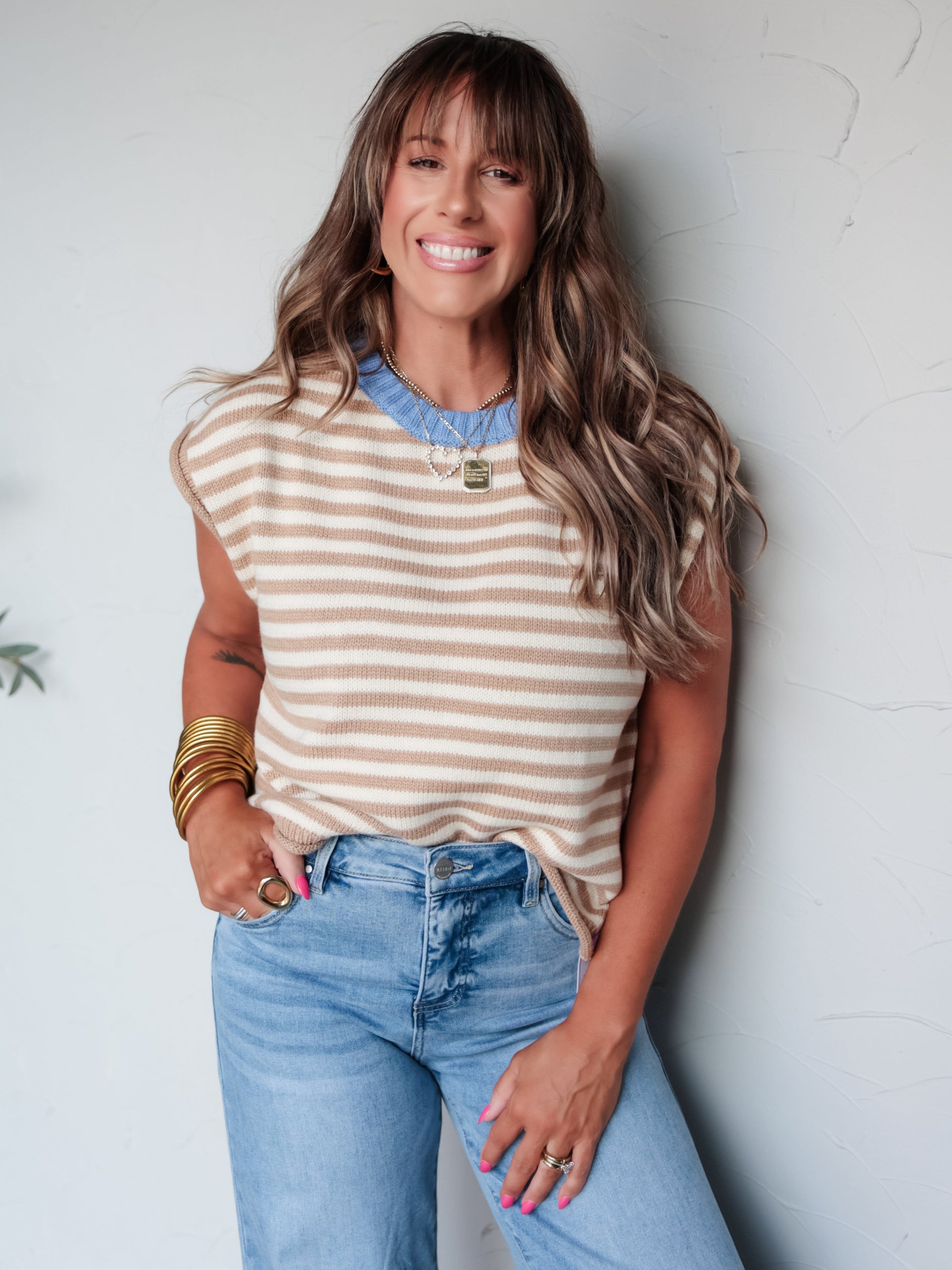 A three-quarter length photograph of a smiling woman with medium-length highlighted hair and bangs posing against a textured white wall. She is wearing a beige and cream horizontal striped sleeveless knit sweater vest made from a 70% polyester and 30% acrylic blend, featuring a contrast light blue crew neckline, paired with light-wash denim jeans and styled with layered pendant necklaces and stacked gold bangles.