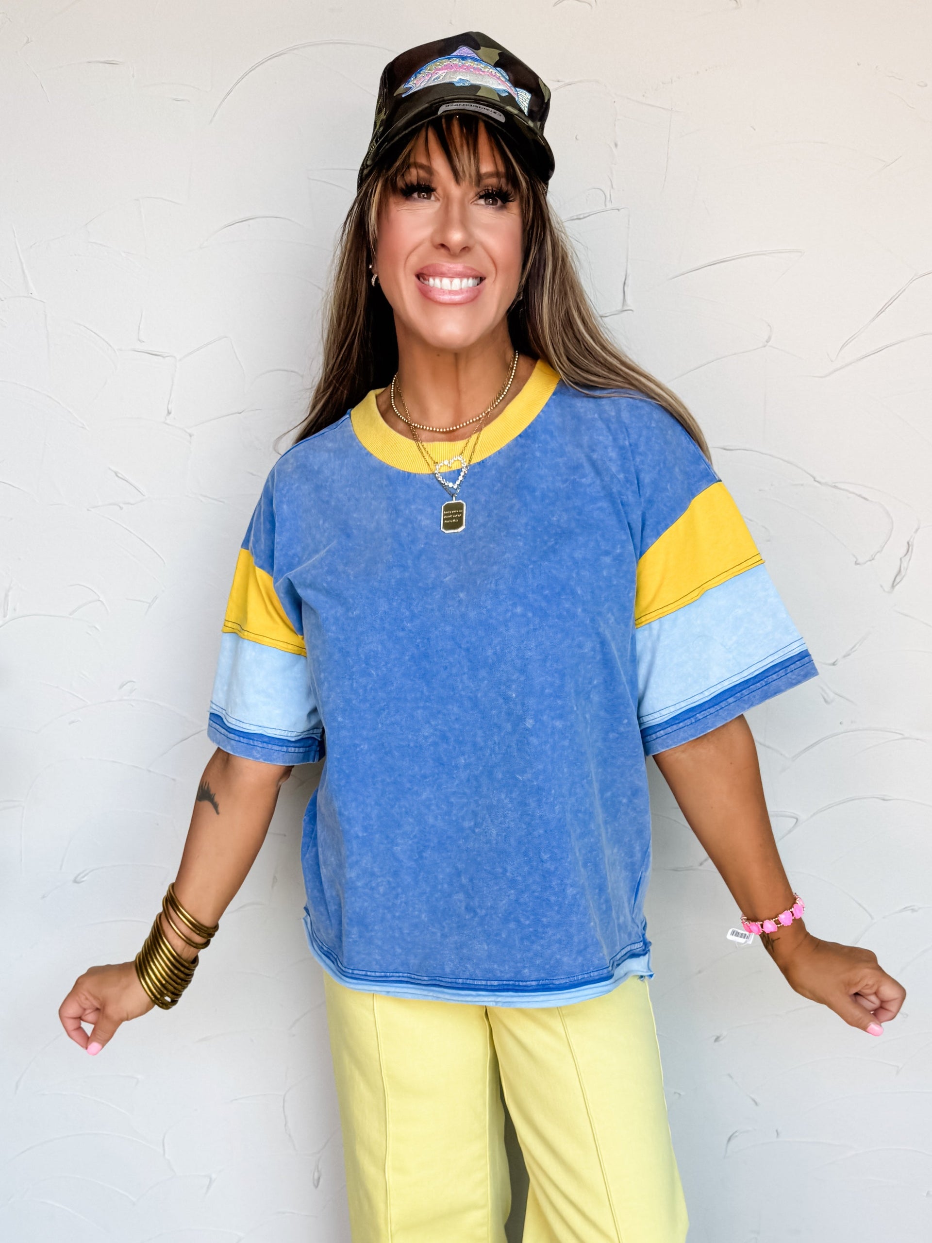 Woman wearing a blue and yellow striped shirt and yellow pants standing against a white wall.