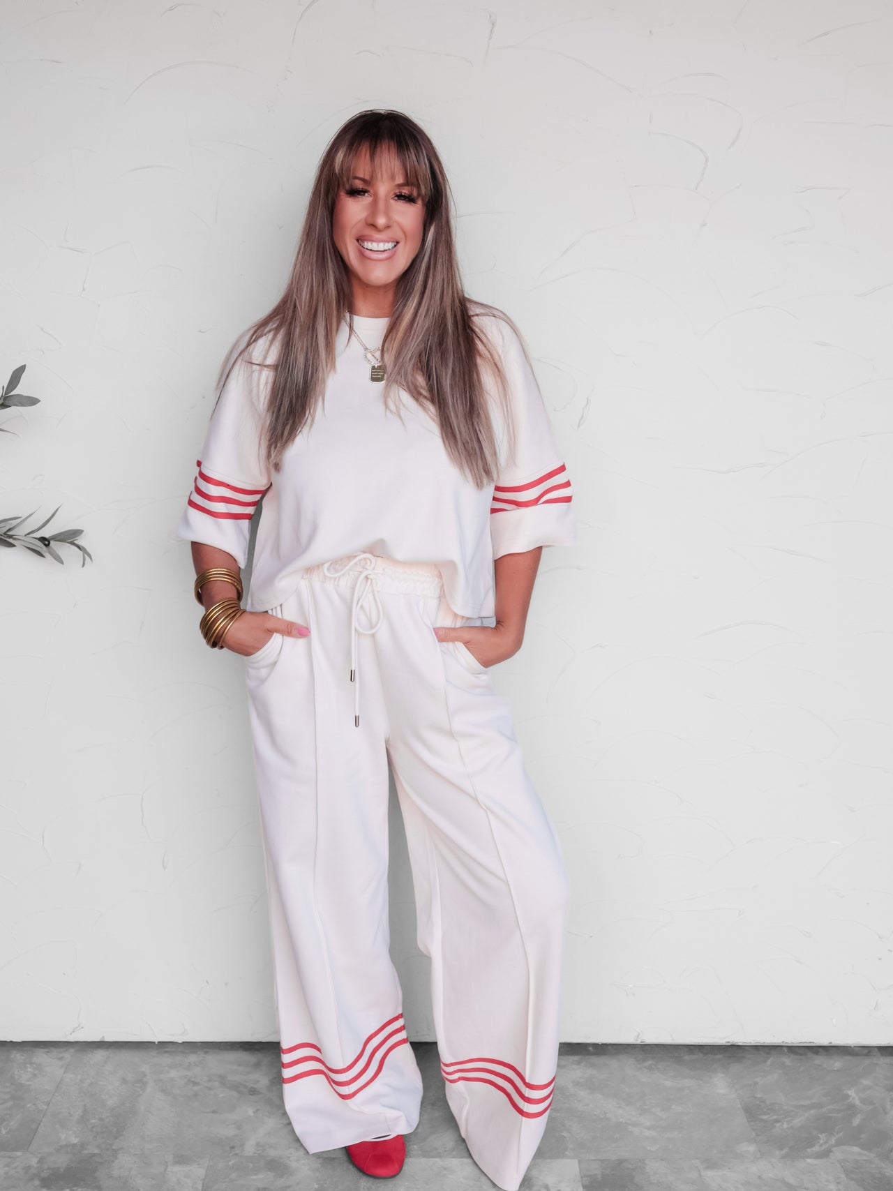 A woman modeling a white two-piece lounge set with red stripe accents on the sleeves and hems, standing against a white wall.