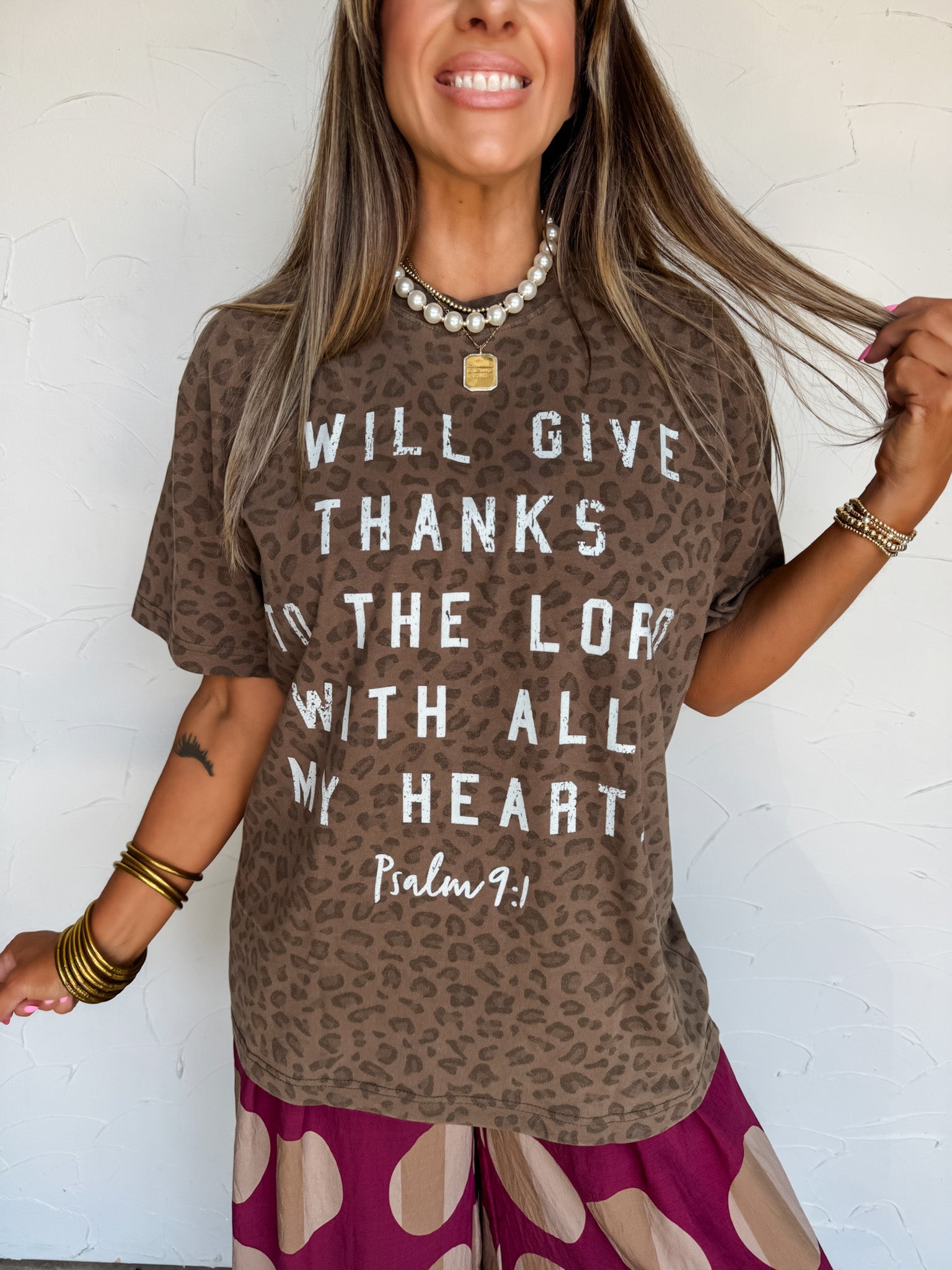 A woman wearing a 100% cotton brown leopard-print graphic t-shirt with white text that reads 'WILL GIVE THANKS TO THE LORD WITH ALL MY HEART Psalm 9:1' paired with purple and beige patterned pants, standing against a white textured wall.