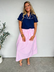 Woman wearing a navy top and pink striped skirt against a white wall.