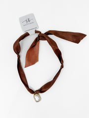 A brown satin ribbon headband with a small gold-toned oval charm, tied in a knot at the top and displayed on a white background with a 'Tiffany Boutique' tag.