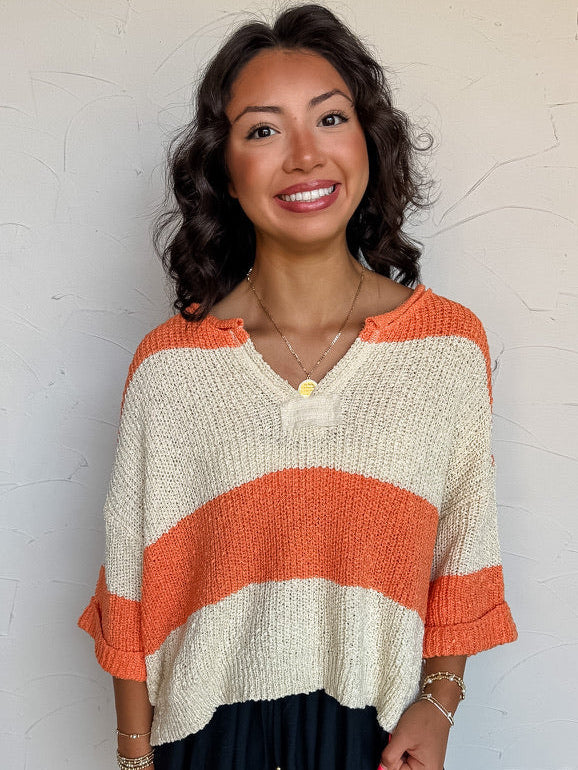 A three-quarter length photograph of a smiling woman with dark wavy hair posing against a textured white wall. She is wearing an open-knit lightweight sweater made from a blend of 54% acrylic, 24% nylon, and 22% polyester, featuring wide horizontal stripes in bright orange and cream, a notched V-neckline, and cuffed elbow-length sleeves, paired with black shorts featuring a lace hem and styled with a gold pendant necklace and gold bracelets.