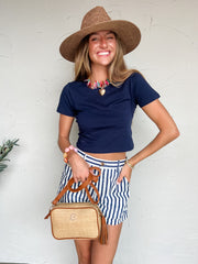 Woman wearing a navy top, striped skirt, and straw hat against a white wall.