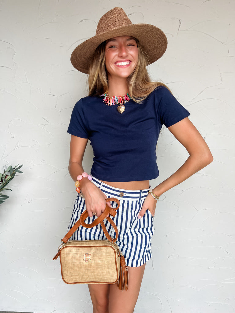 Woman wearing a navy top, striped skirt, and straw hat against a white wall.