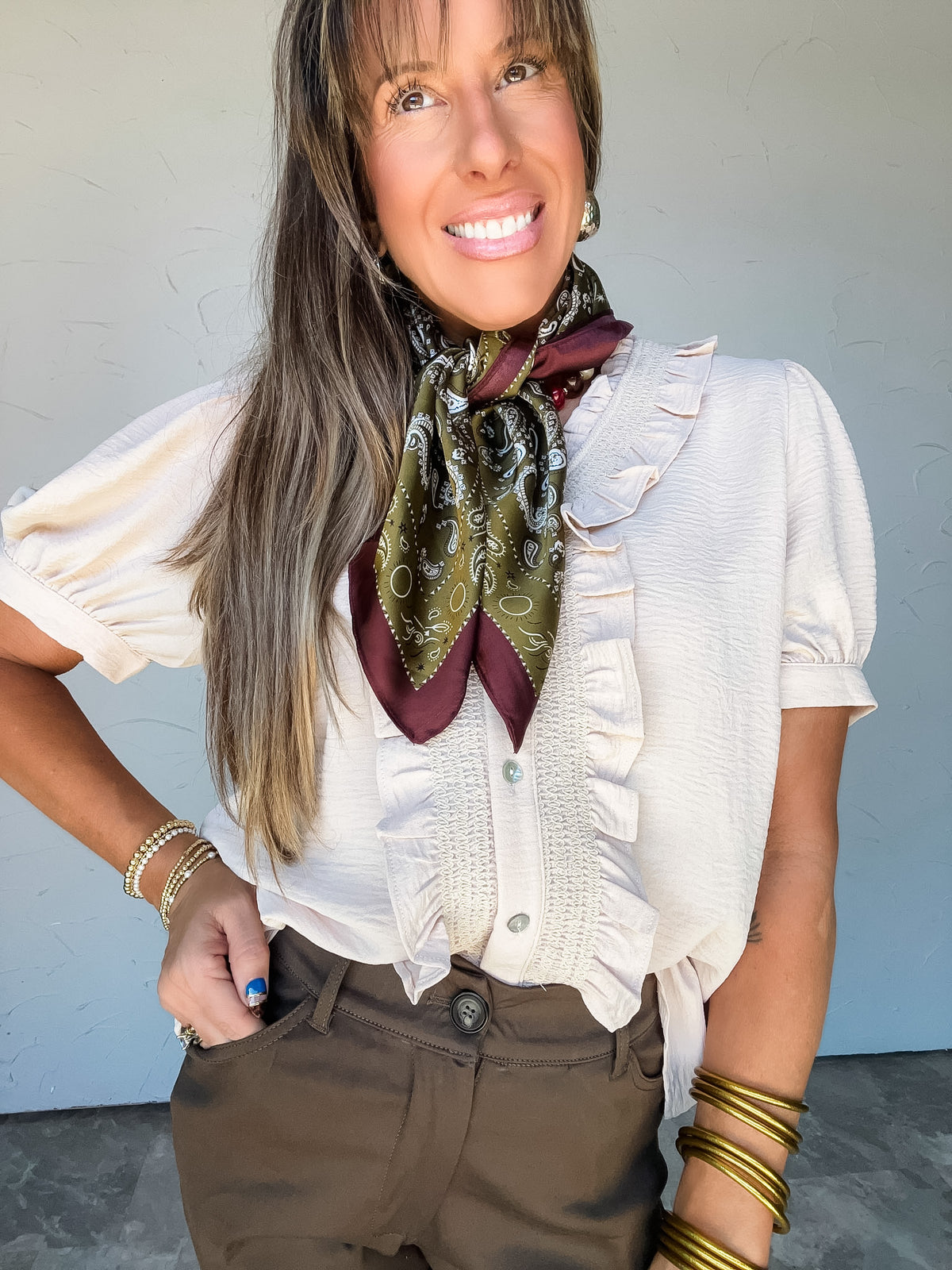 Model wearing the Weekend Whim Ruffle Detail Top in oatmeal by Jodifl, made from 100% polyester, featuring a subtly textured fabric, ruffled V-neckline, button front bordered by vertical ruffles and gathered detailing, and short puff sleeves with fitted cuffs, styled with brown pants and gold-tone jewelry.