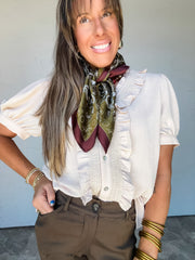 Model wearing the Weekend Whim Ruffle Detail Top in oatmeal by Jodifl, made from 100% polyester, featuring a subtly textured fabric, ruffled V-neckline, button front bordered by vertical ruffles and gathered detailing, and short puff sleeves with fitted cuffs, styled with brown pants and gold-tone jewelry.