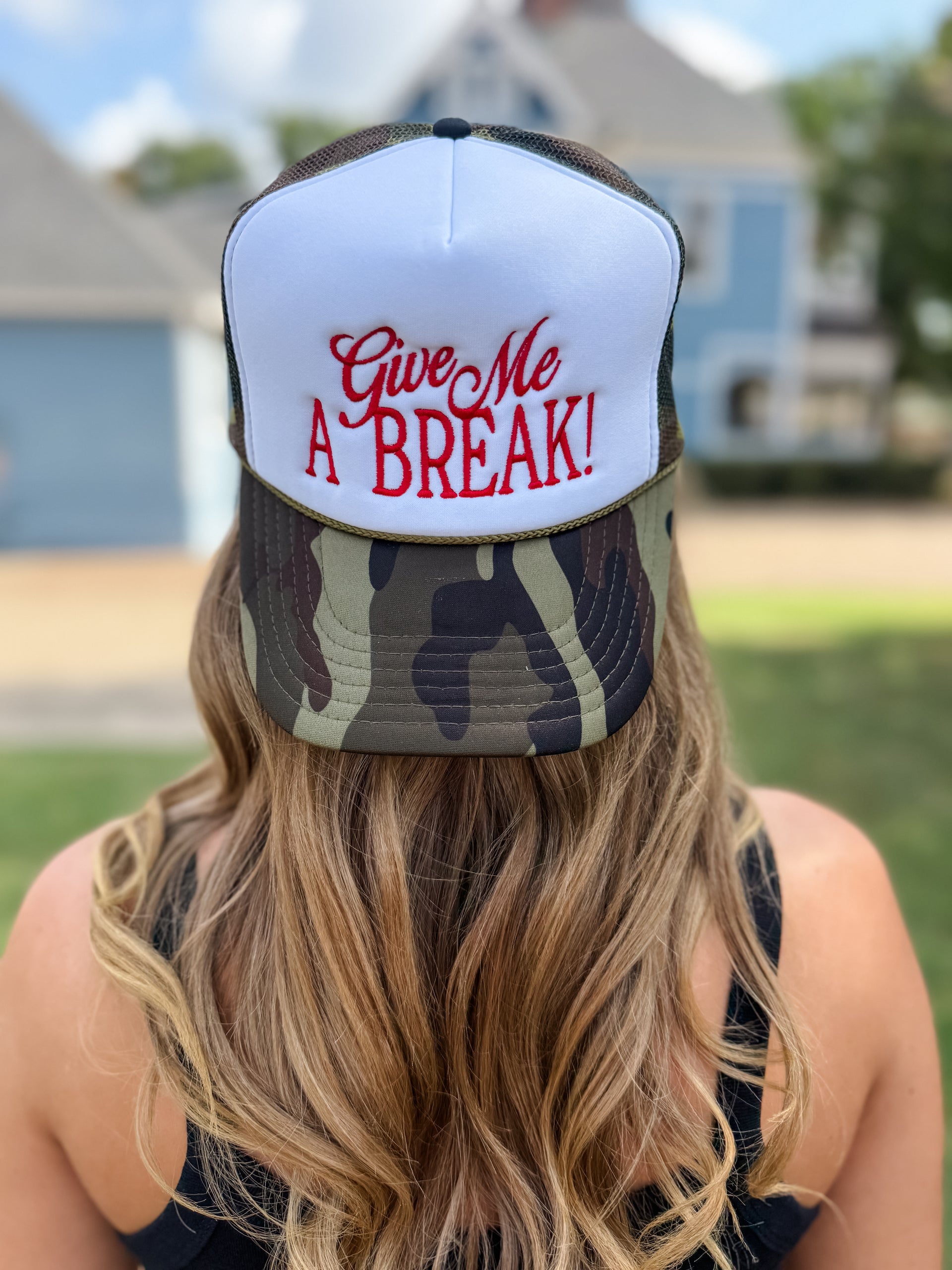 A woman viewed from behind wearing a white and camouflage trucker hat with red embroidered text that reads 'Give Me A BREAK!' against an outdoor residential background.