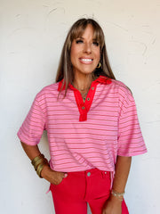 A waist-up photograph of a smiling woman with straight light-brown hair and front bangs posing with one hand in her pocket against a textured white wall. She is wearing a short-sleeved polo top made from a 65% polyester, 30% cotton, and 5% spandex blend, featuring light pink and red horizontal stripes, wide elbow-length sleeves, and a vibrant red collar and button placket, front-tucked into hot pink pants and styled with stacked gold bangles, gold hoop earrings, and a layered gold chain necklace.