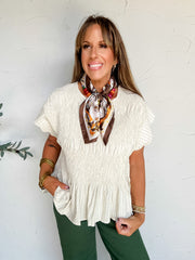 A portrait photograph of a smiling woman with long light-brown hair and bangs posing against a textured off-white wall next to a potted olive plant with her hand in her pocket. She is wearing a short-sleeved cream pinstriped blouse made from a 55% linen and 45% rayon blend, featuring a smocked bodice, flutter cap sleeves, and a peplum hem, paired with forest green pants, styled with a brown equestrian-patterned silk neck scarf over a colorful beaded necklace, stacked gold bangles, and beaded bracelets.