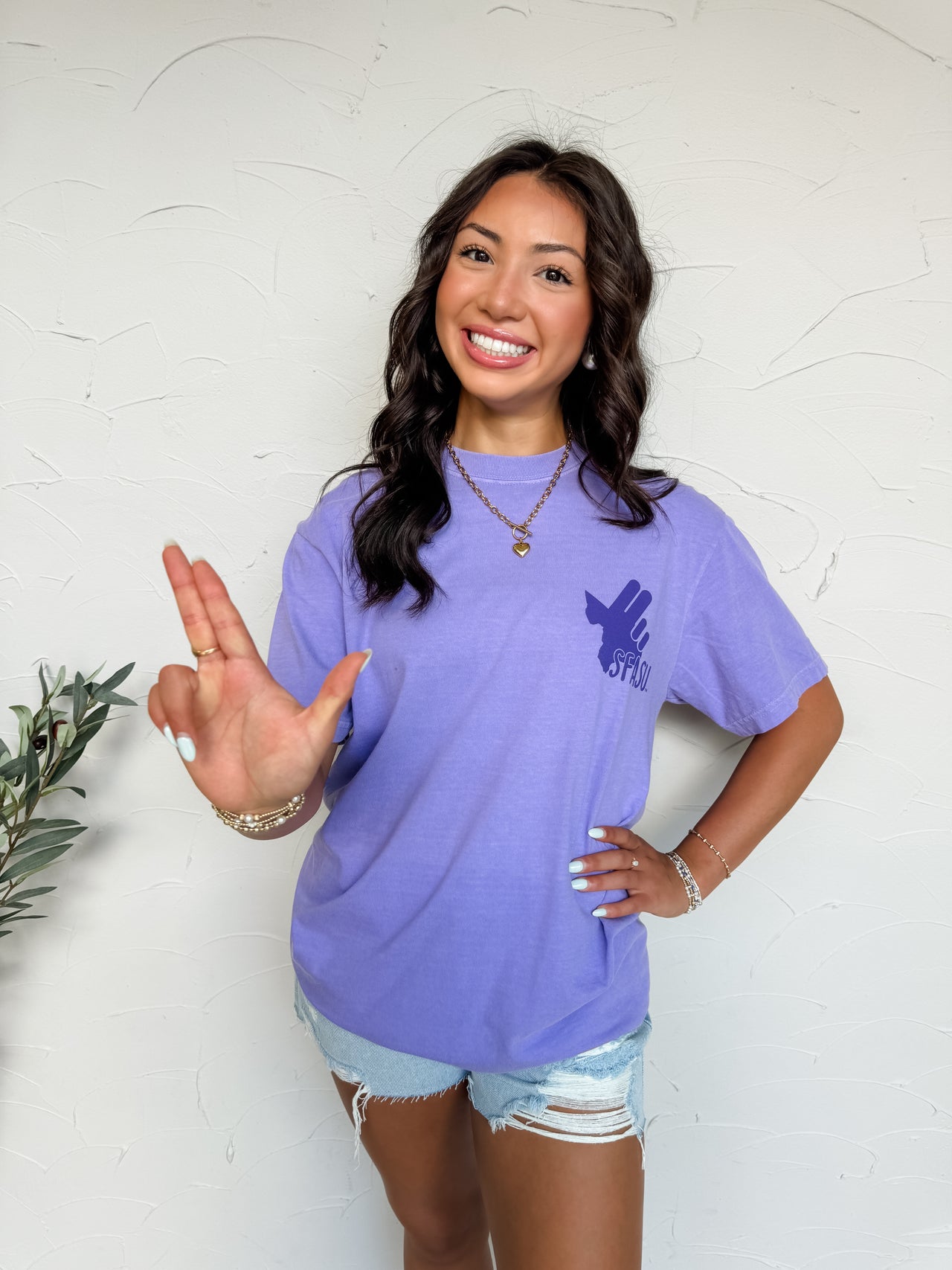 Woman wearing a purple t-shirt with a logo, standing against a white wall.