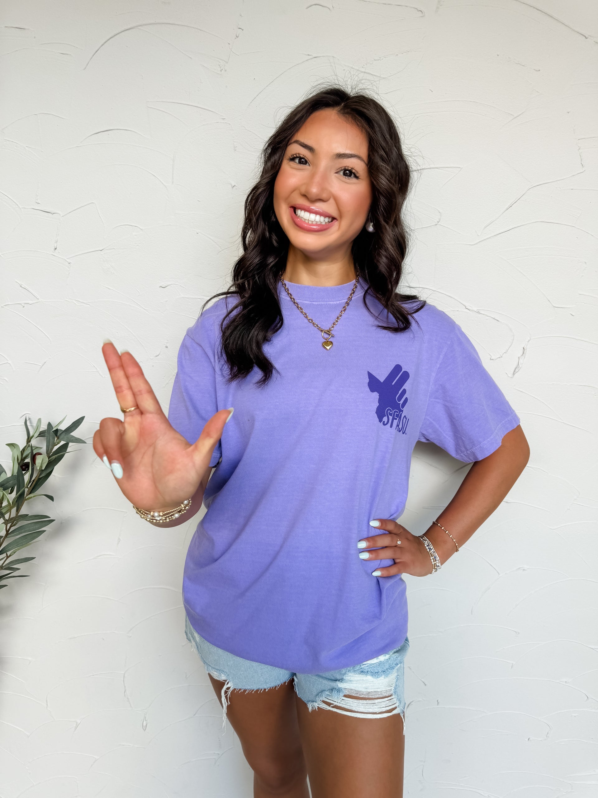 Woman wearing a purple t-shirt with a logo, standing against a white wall.