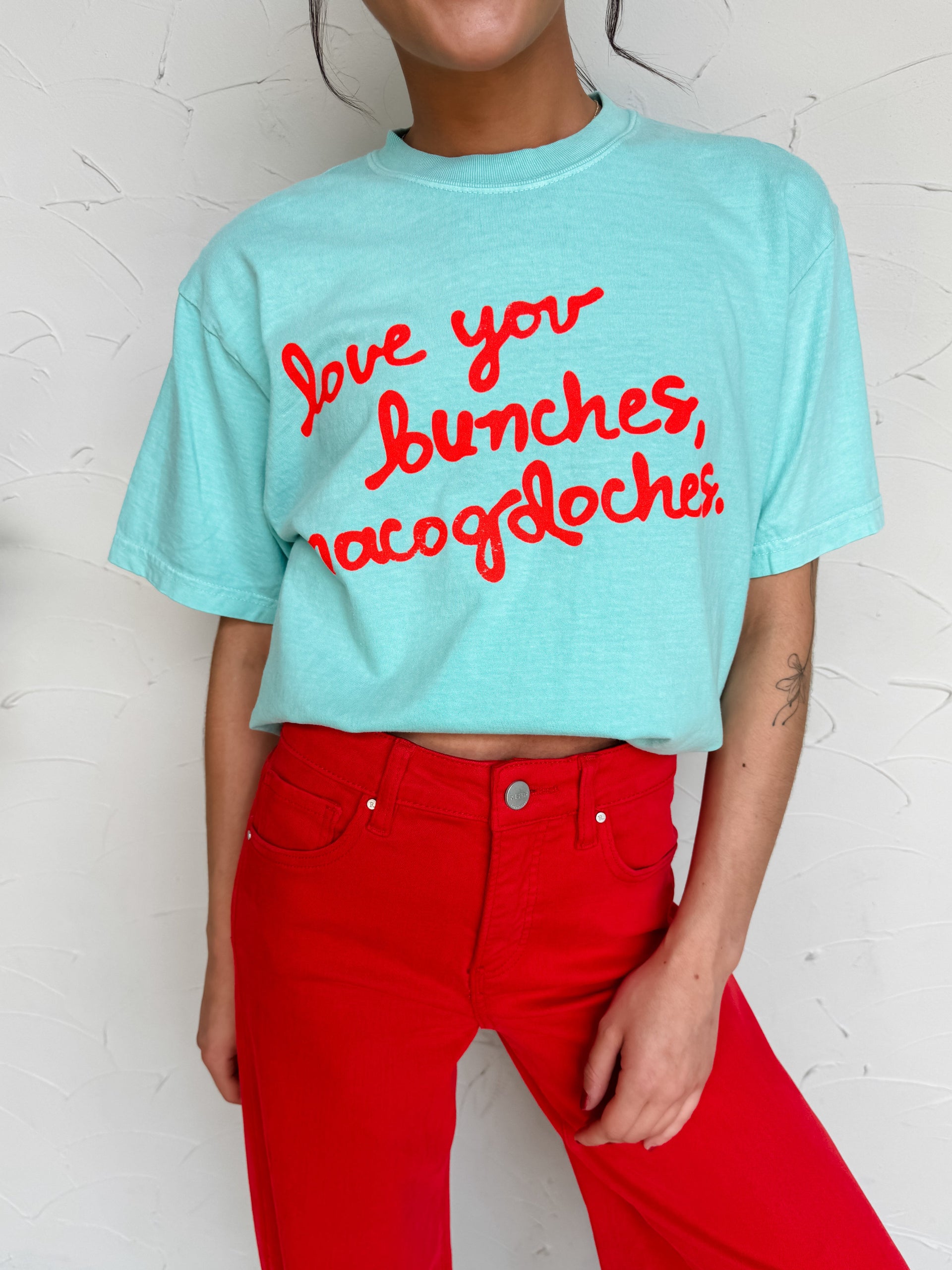 A close-up view of a person standing against a textured off-white wall, wearing a light aqua blue graphic t-shirt made from 100% cotton. The shirt features the phrase "love you bunches, nacogdoches." printed across the front in a vibrant red cursive script. The top is styled slightly tucked under to create a cropped look and is paired with high-waisted, bright red denim pants featuring standard five-pocket styling and a silver button closure.