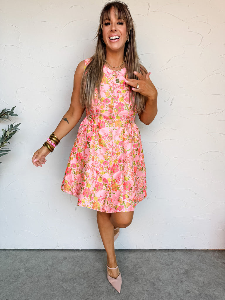 A smiling woman with long brown hair stands against an off-white wall, wearing a sleeveless pink floral mini dress made from an 85% polyester and 15% nylon blend. The structured jacquard dress features a round neckline, a full button-down front, a fitted waist, and a flared skirt covered in a vibrant pink, green, and orange floral pattern. She is accessorized with stacked gold bangles, layered gold necklaces, and statement rings.