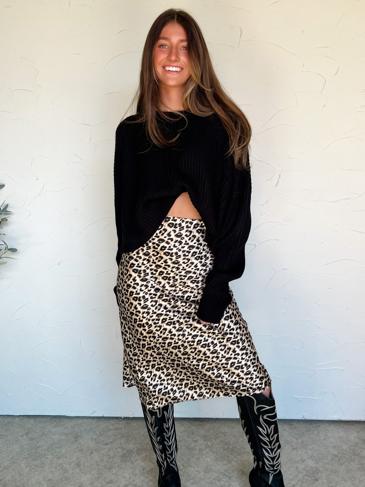 Woman wearing a black sweater, leopard print skirt, and black knee-high boots against a white wall.