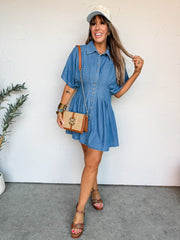 A smiling woman with long hair leans against an off-white wall, wearing a short-sleeve medium-wash denim mini dress made from 100% cotton. The dress features a classic point collar, a full button-down front, and structural stitched pleats at the waist that create a tailored, cinched silhouette. She is accessorized with a light-colored trucker hat, stacked gold bangles with blue rhinestone accents, and a straw shoulder bag with brown leather trim and a gold logo emblem.