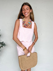 Woman in a pink dress with a colorful necklace holding a straw bag against a white wall.