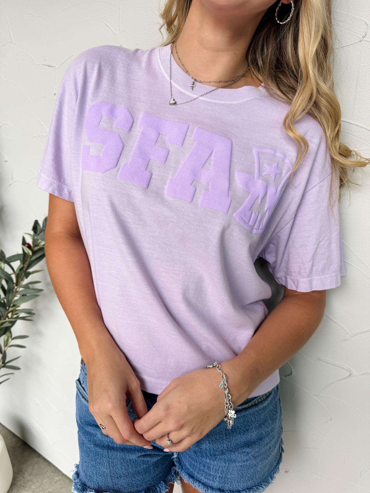 Person wearing a light purple t-shirt with 'SFA' printed on it, standing against a white wall.