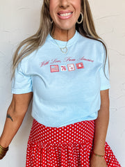 Woman wearing a light blue shirt with "with love, from america" and patriotic stamp graphics, paired with a red polka dot skirt against a white background.