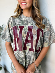 A close-up of woman wearing a short-sleeve camouflage graphic t-shirt with the letters 'NAC' printed in dark red across the chest, posing against a white wall.