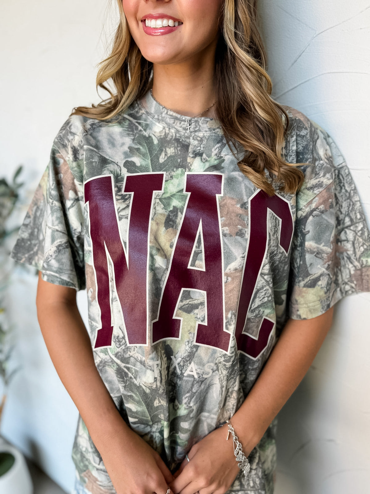 A close-up of woman wearing a short-sleeve camouflage graphic t-shirt with the letters 'NAC' printed in dark red across the chest, posing against a white wall.