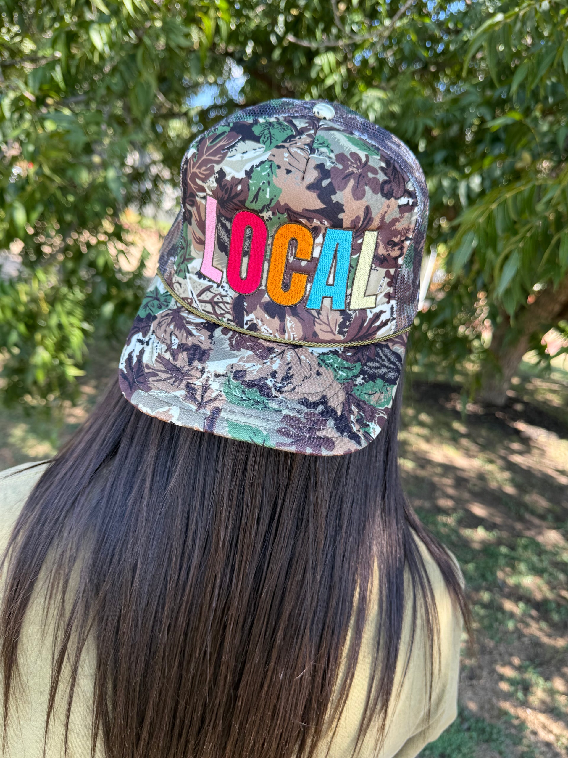 A person outdoors wearing a camouflage baseball cap with the word 'LOCAL' embroidered across the front in colorful letters.