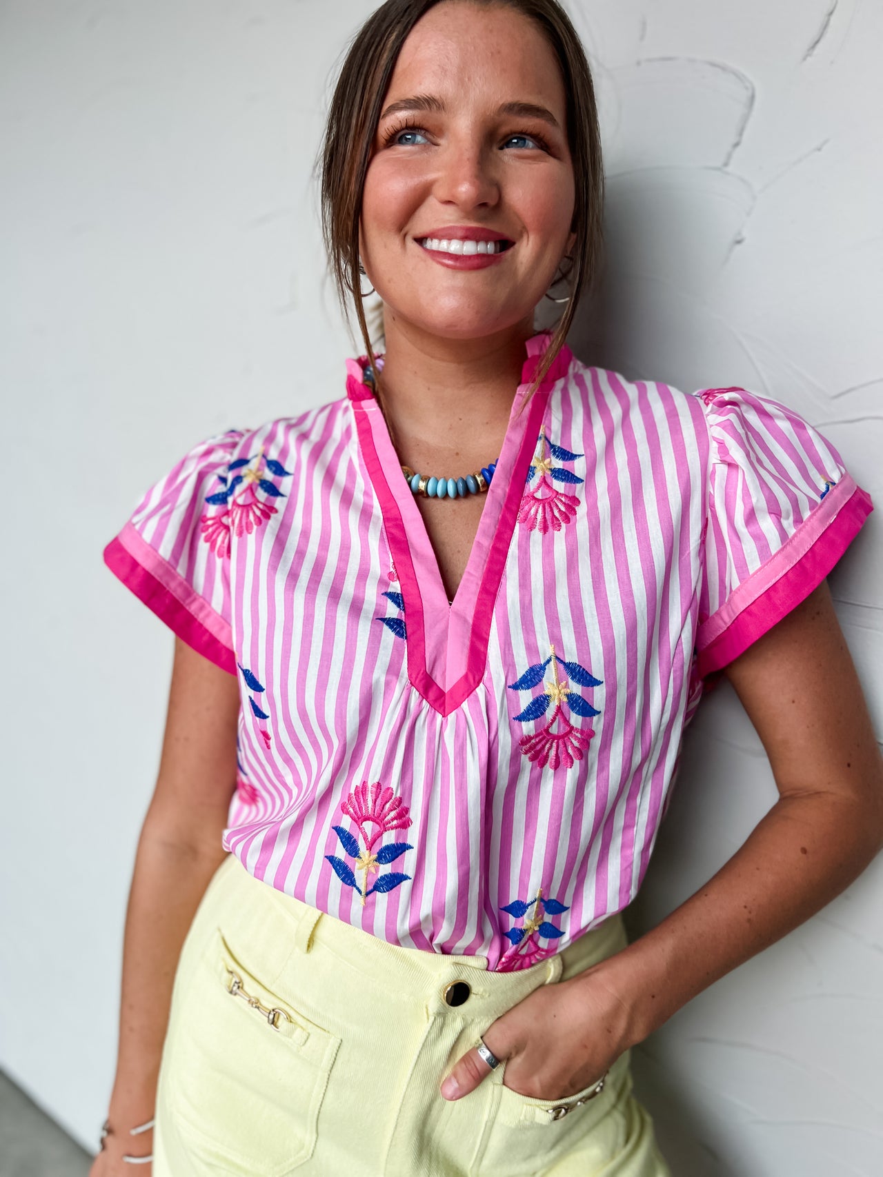 Woman wearing a pink and white striped blouse with floral embroidery against a white wall.