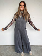 Woman wearing a gray jumpsuit with long sleeves and tattoos, standing against a white wall.