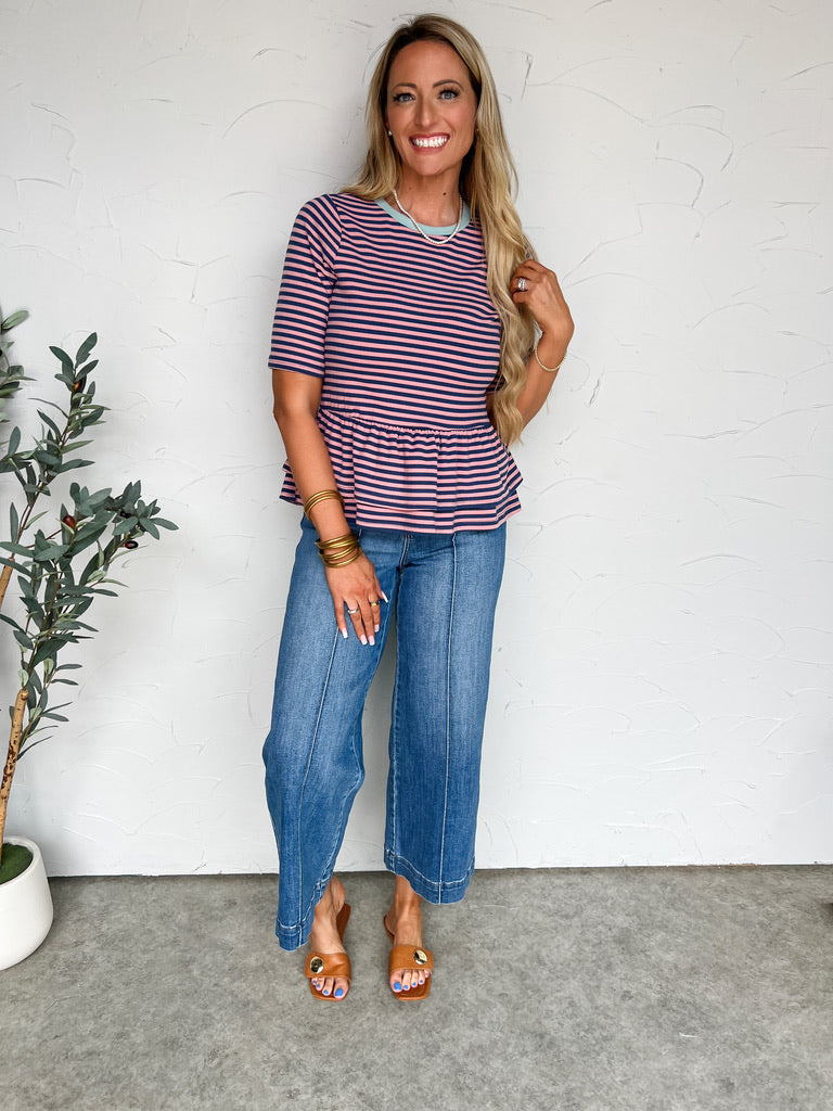 Woman wearing a striped shirt and jeans standing against a white wall with a plant to her left.