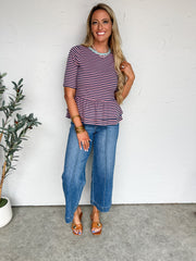 Woman wearing a striped shirt and jeans standing against a white wall with a plant to her left.