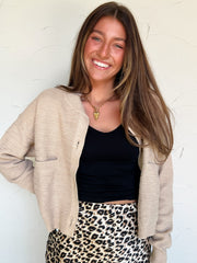 Woman wearing a beige cardigan, black top, and leopard print skirt against a white wall.