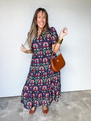 Model wearing the Made With Love Tiered Printed Midi Dress, made from 40% rayon, 30% polyester, and 30% Tencel, featuring pink, cream, and rust floral motifs on a dark teal background, a notched neckline, short puff sleeves, a gathered waist, and a flowing tiered skirt, styled with a cognac handbag and brown shoes.