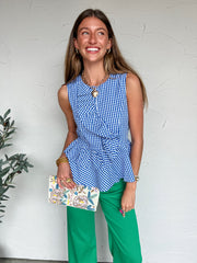 Woman wearing a blue and white checkered top and green pants, holding a floral clutch against a white wall.
